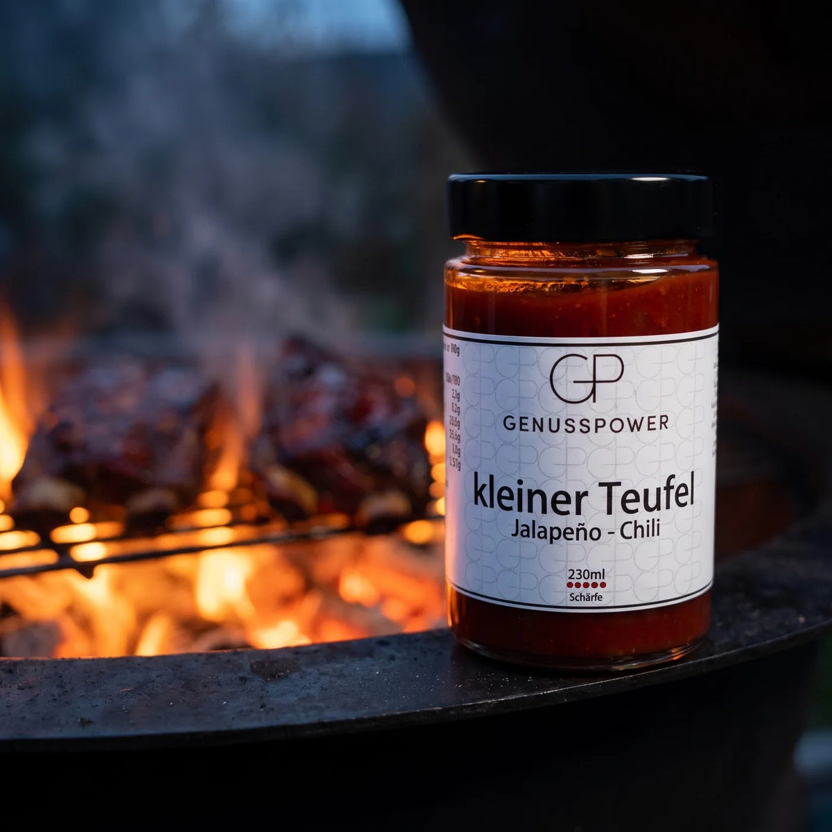 Glas kleiner Teufel auf dem Stahlrand eines Holzkohlegrills, dahinter Flammen und Rippchen