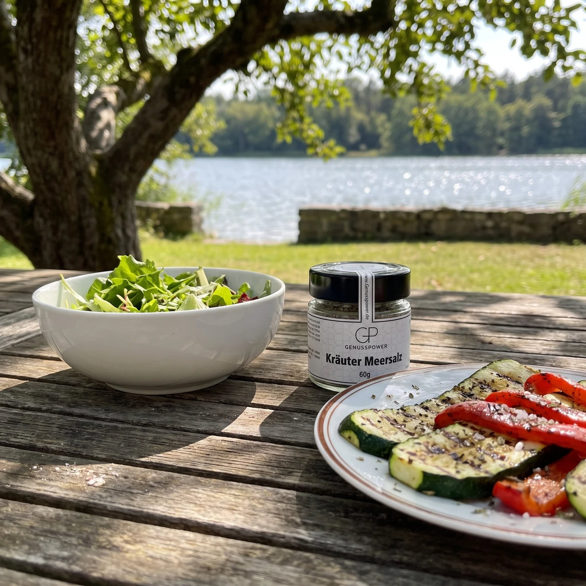 Glas Kräuter Meersalz von Genusspower auf einem Gartentisch am See, daneben eine Schüssel Salat und ein Teller mit gegrilltem Gemüse