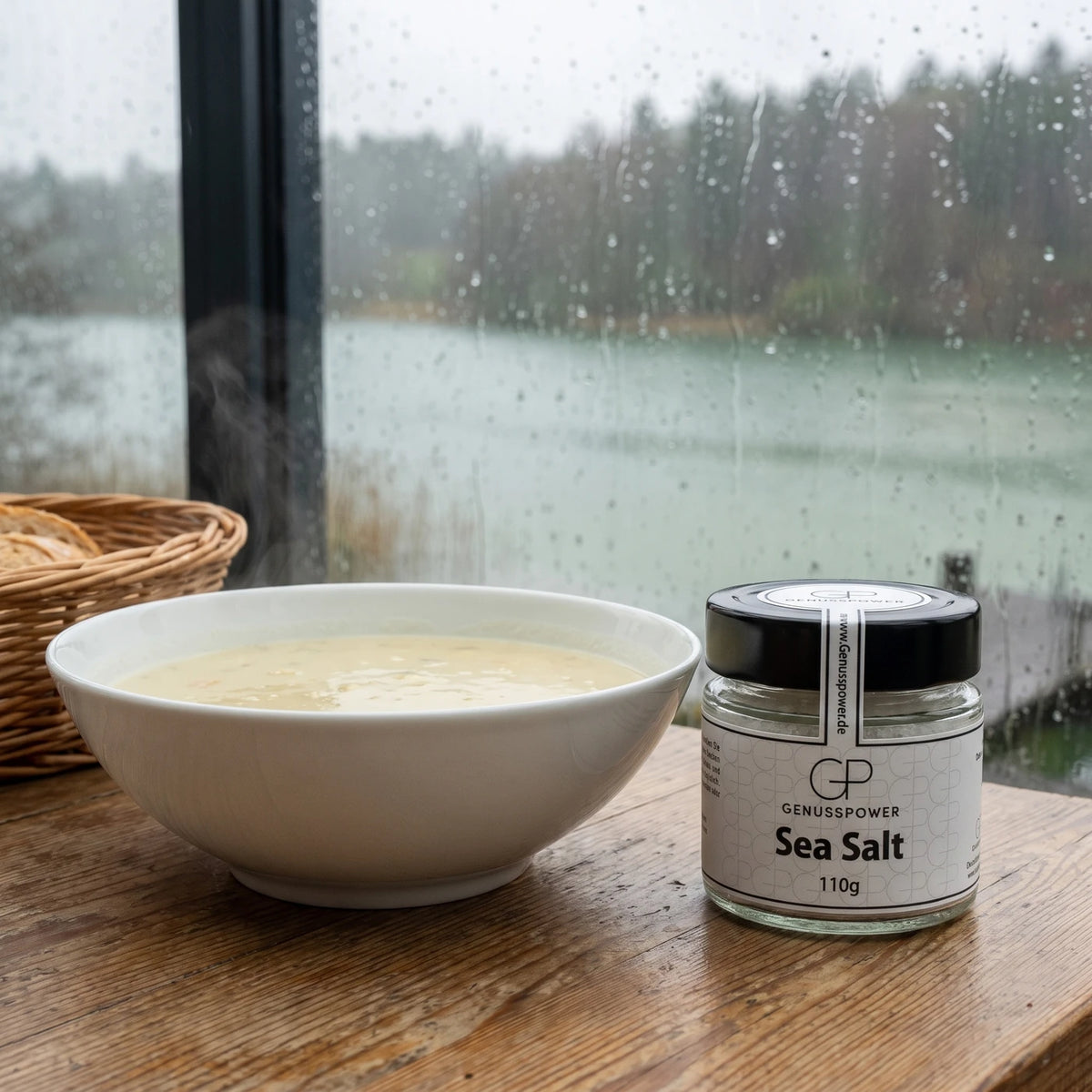 Glas Sea Salt von Genusspower auf einem Tisch im Wintergarten am See, daneben eine Schüssel Kartoffelsuppe und ein Brotkorb, Regen am Fenster