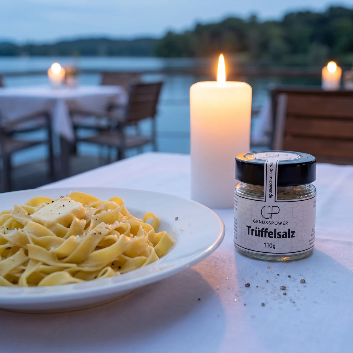 Glas Trüffelsalz von Genusspower auf einem weiß gedeckten Tisch auf einer Terrasse am See in der Dämmerung, daneben ein Teller Tagliatelle und eine Kerze